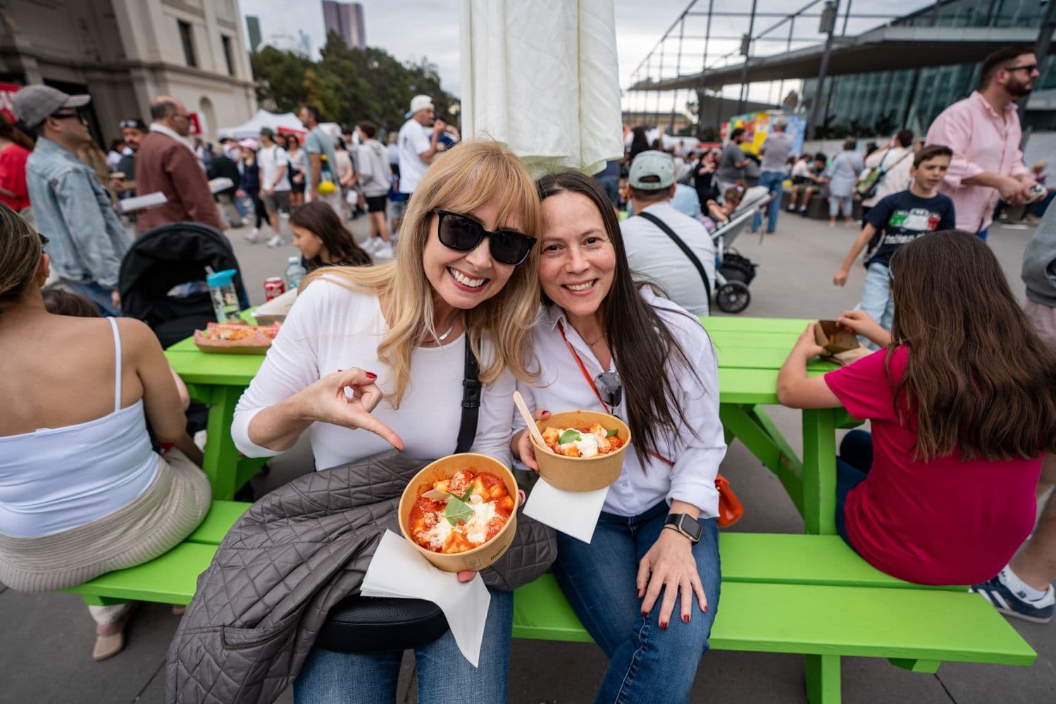 Two women smiling at outdoor dining table with Italian pasta dishes at Melbourne Italian Festa