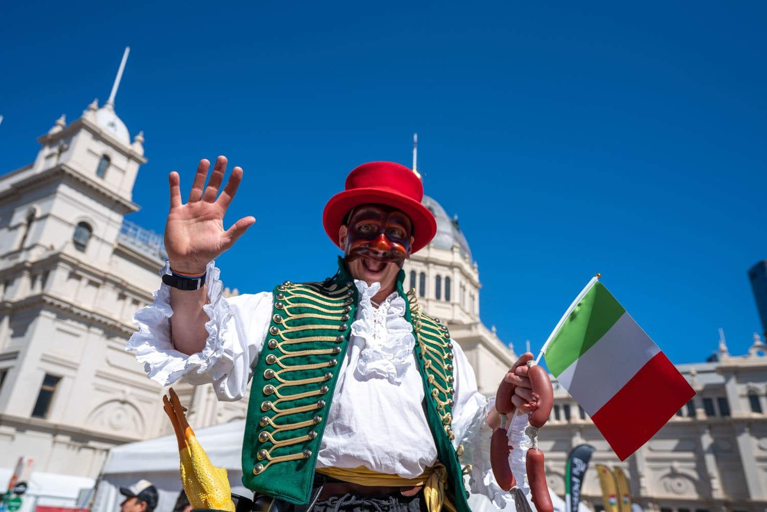 Performer in red hat and ornate green military jacket waves holding Italian flag at Melbourne Festa