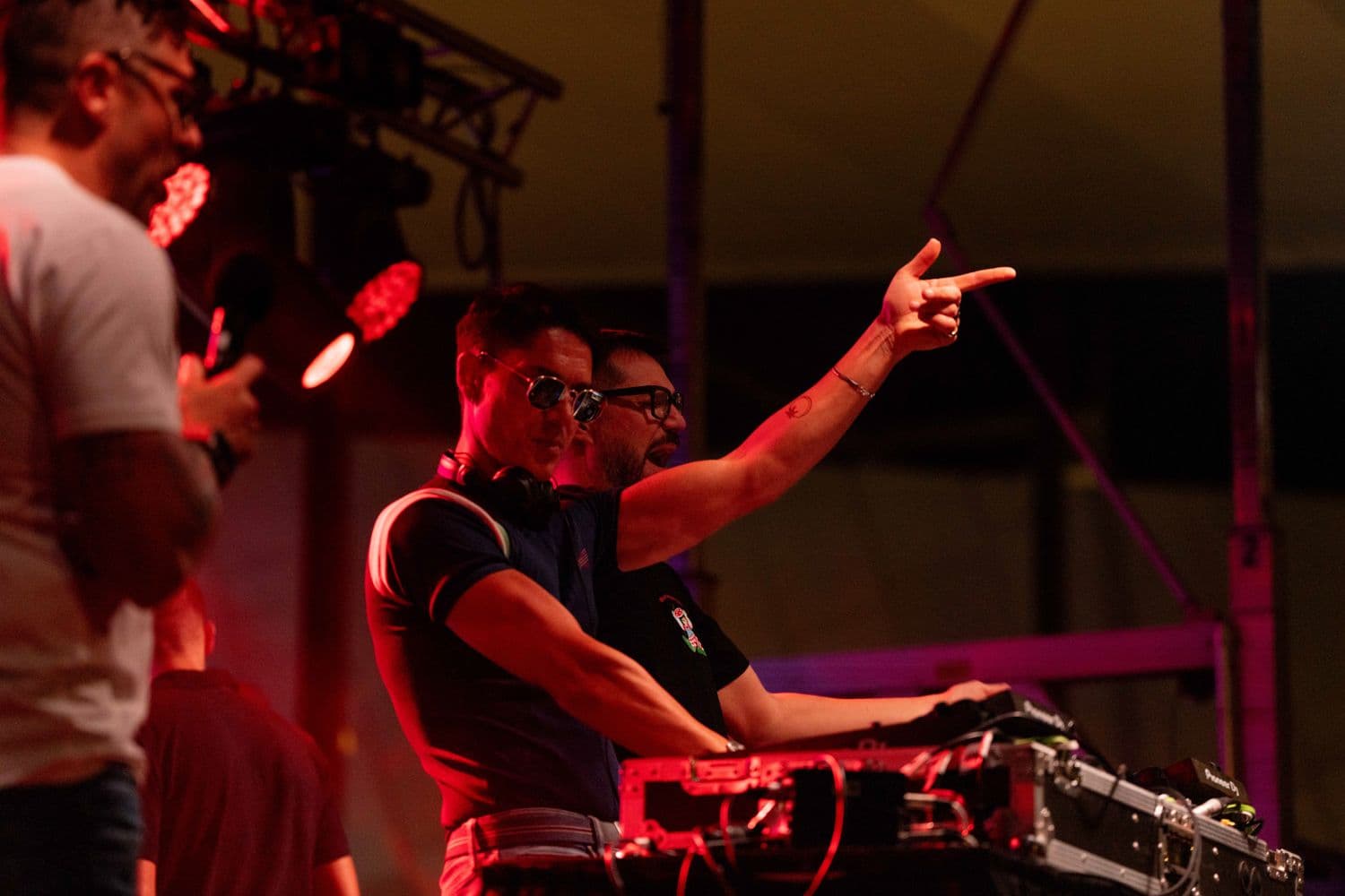 DJ in glasses and black shirt pointing to crowd at illuminated stage with red lighting