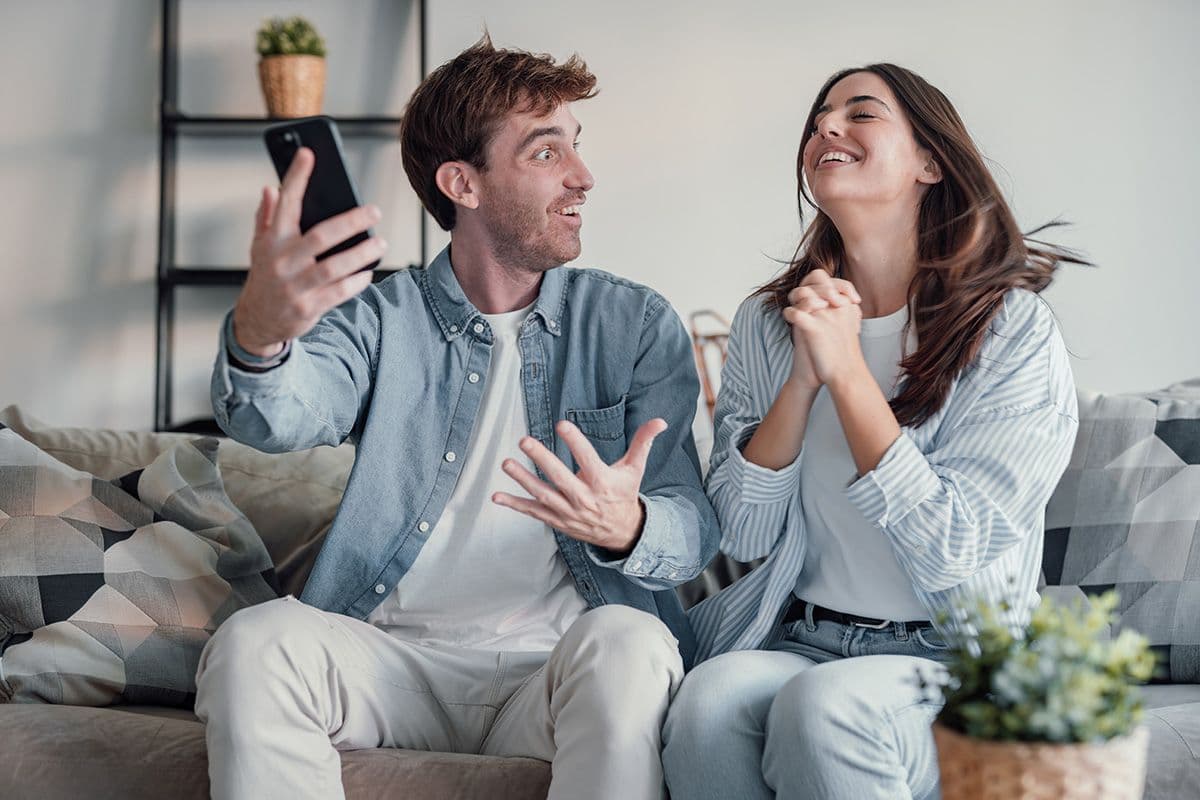 Two people on a sofa smiling while taking a selfie with a smartphone